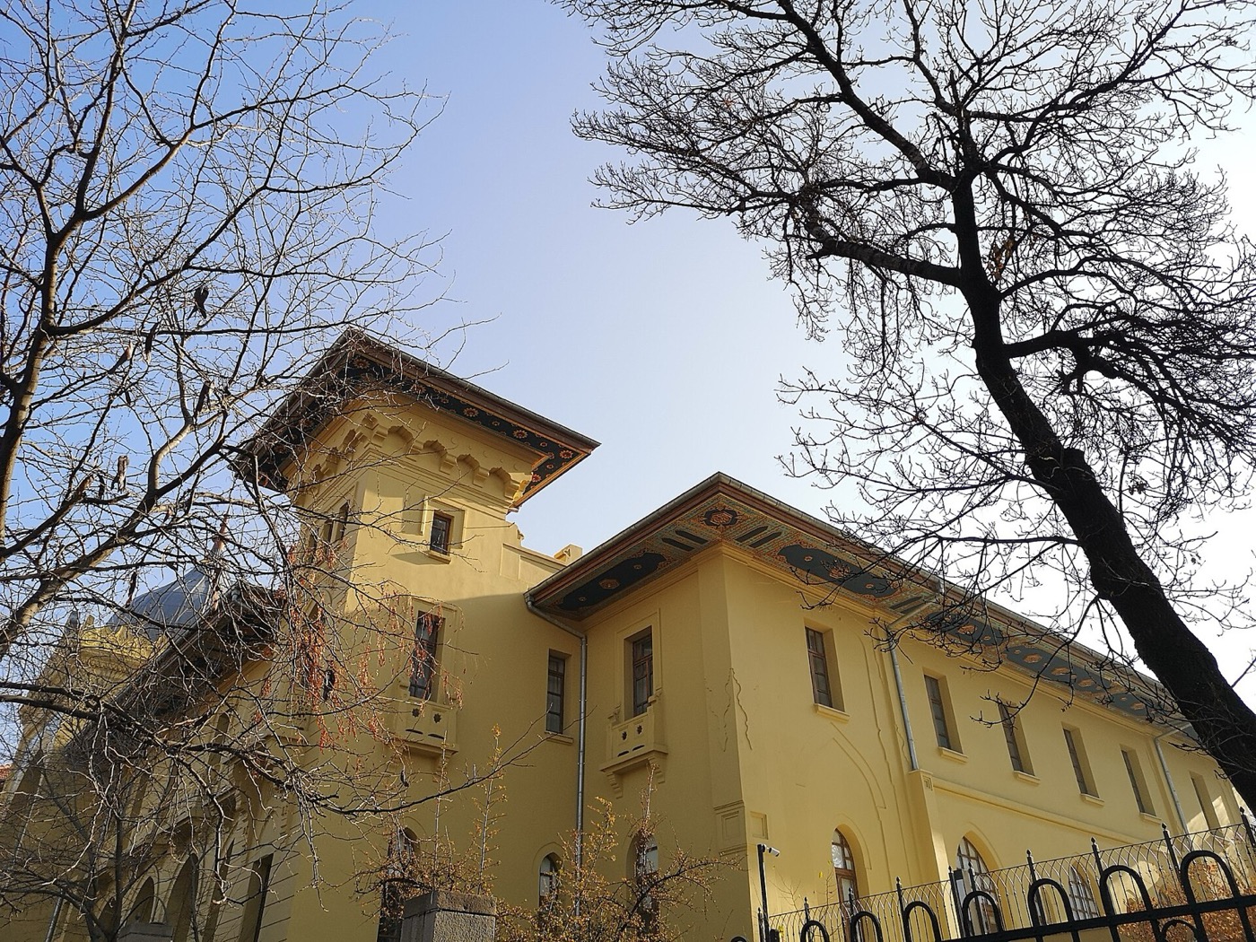 The yellow side wing and corner tower of Ankara Palas behind bare winter trees and an iron fence