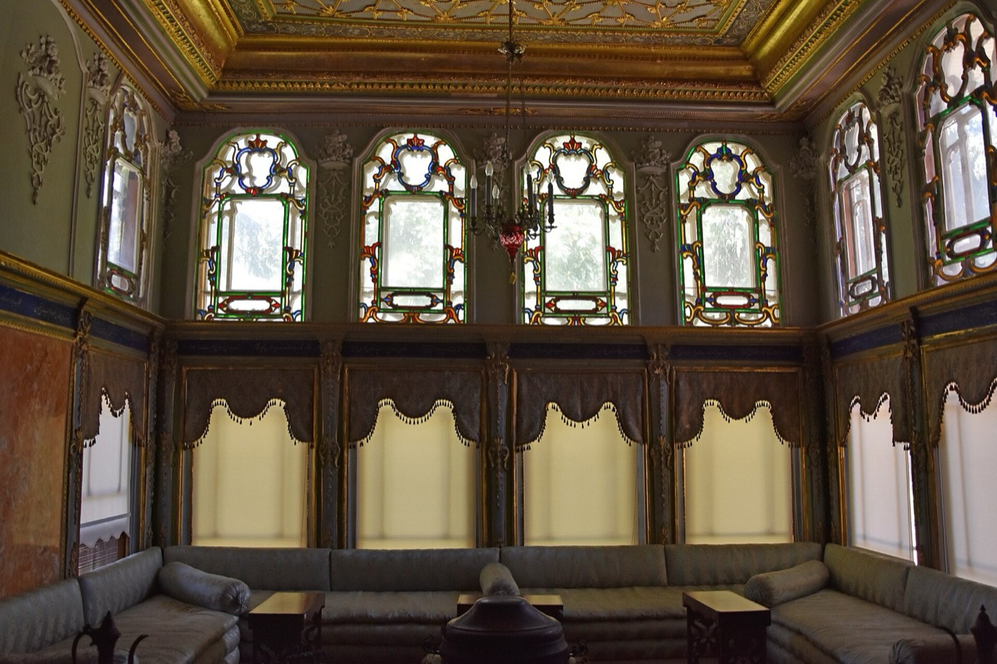 A room in Aynalıkavak Pavilion with a long low divan running along the walls, under a row of arched stained-glass windows and a gilded, painted ceiling