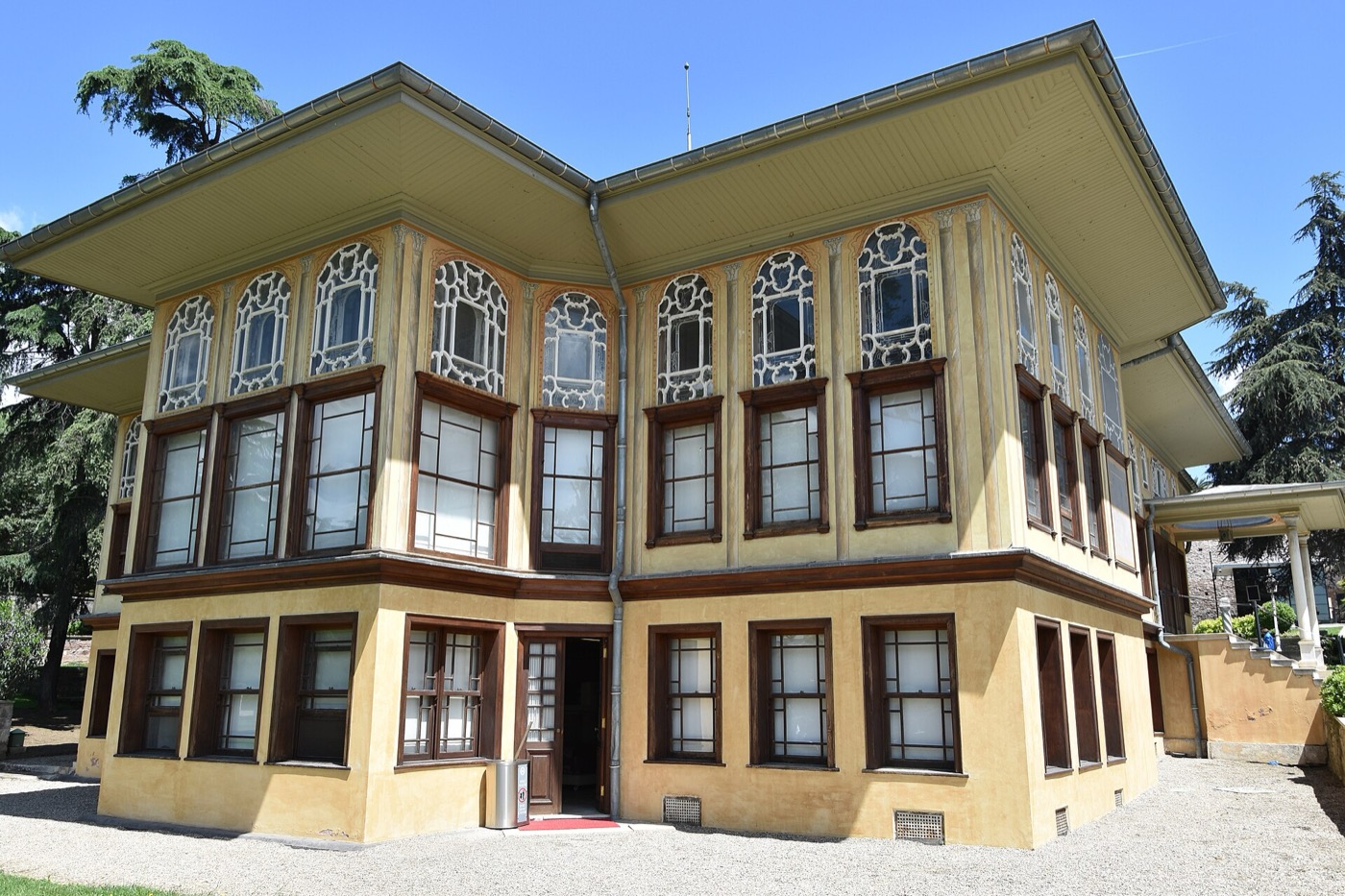 Aynalıkavak Pavilion seen from its garden: a two-storey ochre building with latticed upper windows under wide eaves, on a gravel forecourt