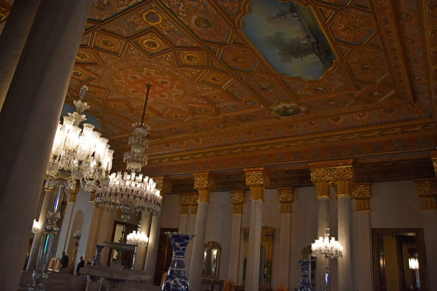 A hall in Beylerbeyi Palace with white columns topped by gilded capitals, crystal chandeliers and a coffered ceiling with painted seascapes