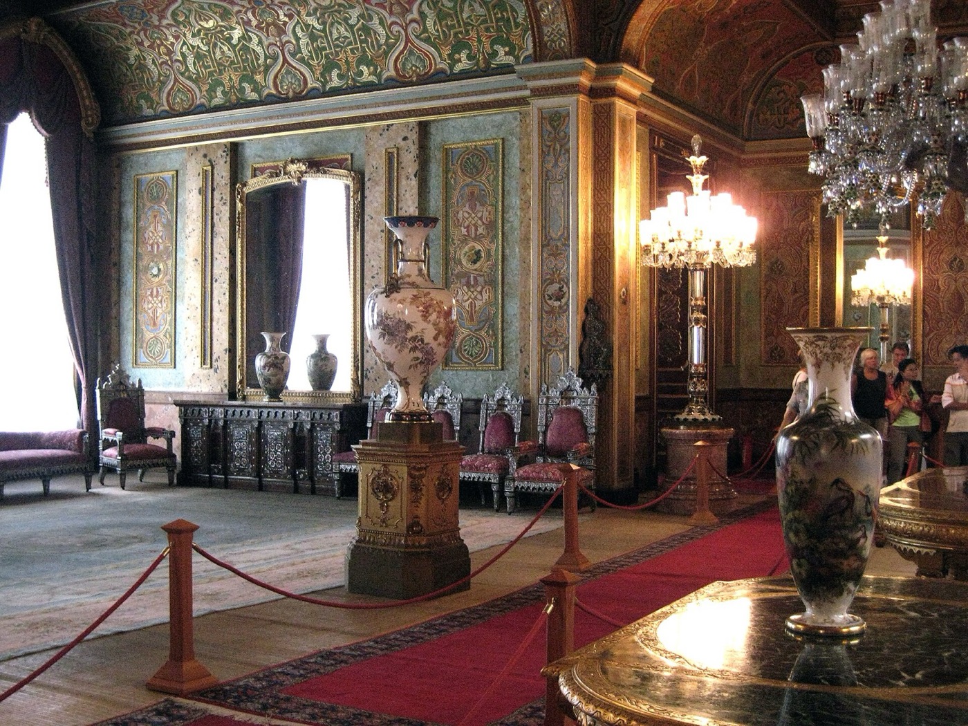 A room in Beylerbeyi Palace with painted wall panels, calligraphy on the ceiling, tall porcelain vases, crystal candelabra and a roped-off visitor path on a red runner