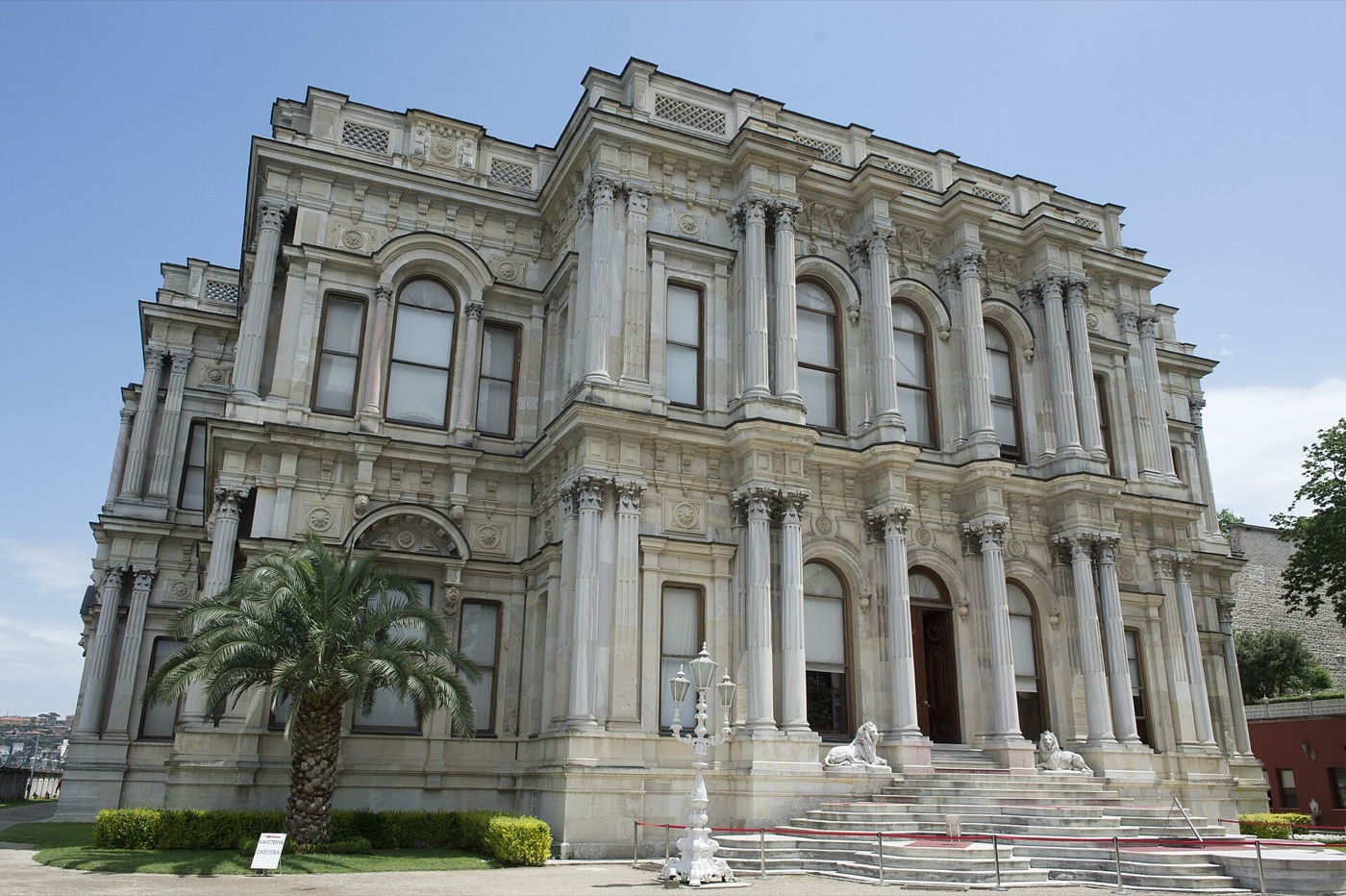 The marble facade of the main palace building at Beylerbeyi, with a flight of steps flanked by two reclining lion statues and a palm tree in front