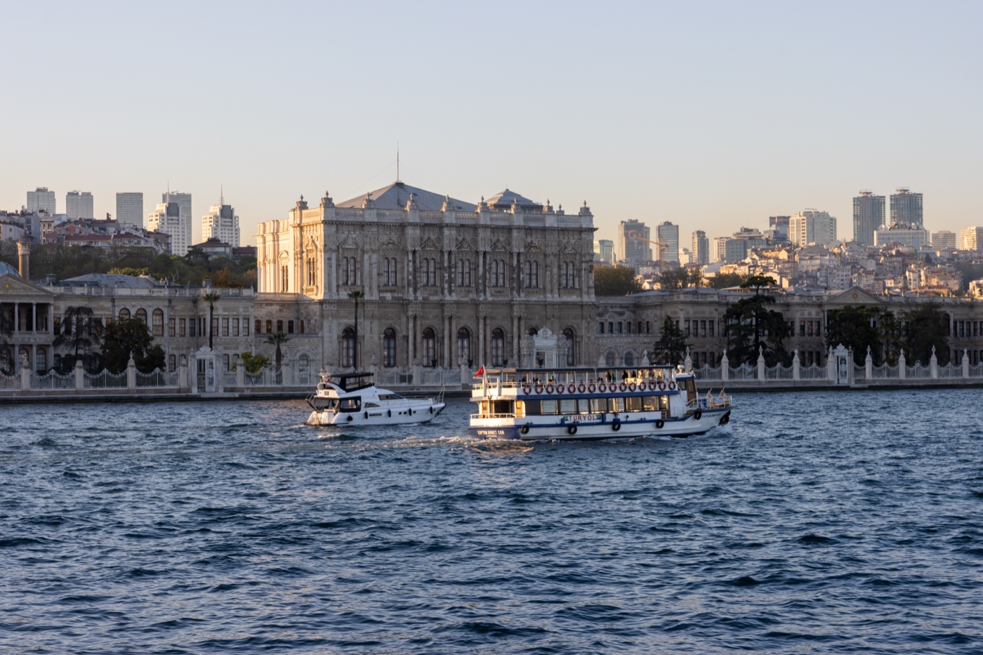Dolmabahçe Palace seen from the Bosphorus: a tall central block between lower wings, behind a white waterfront railing, with a yacht and a passenger boat on the water in front