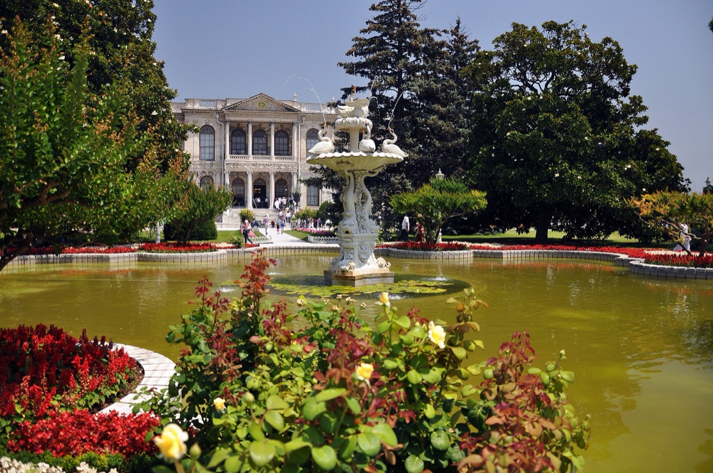 The Swan Fountain in the garden of Dolmabahçe Palace: a tiered marble fountain in a round pool, with flowerbeds in front and the palace façade beyond the trees