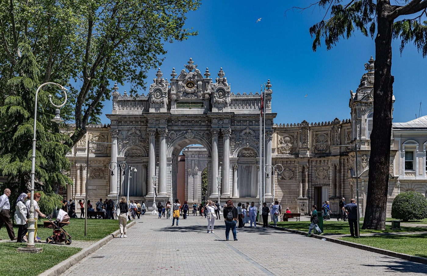 The Treasury Gate of Dolmabahçe Palace seen from inside the grounds: a carved white marble arch with paired columns, at the end of a paved path busy with visitors