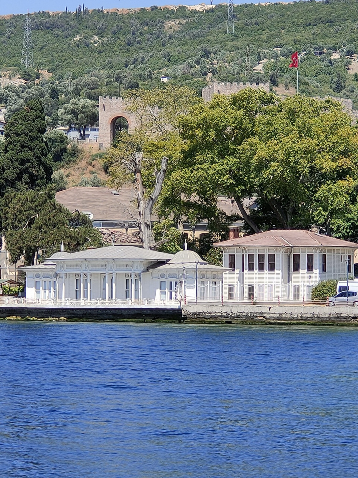 The white timber Kaiser Wilhelm pavilion on the Hereke shore seen from the sea, with the wooded hillside and castle walls above