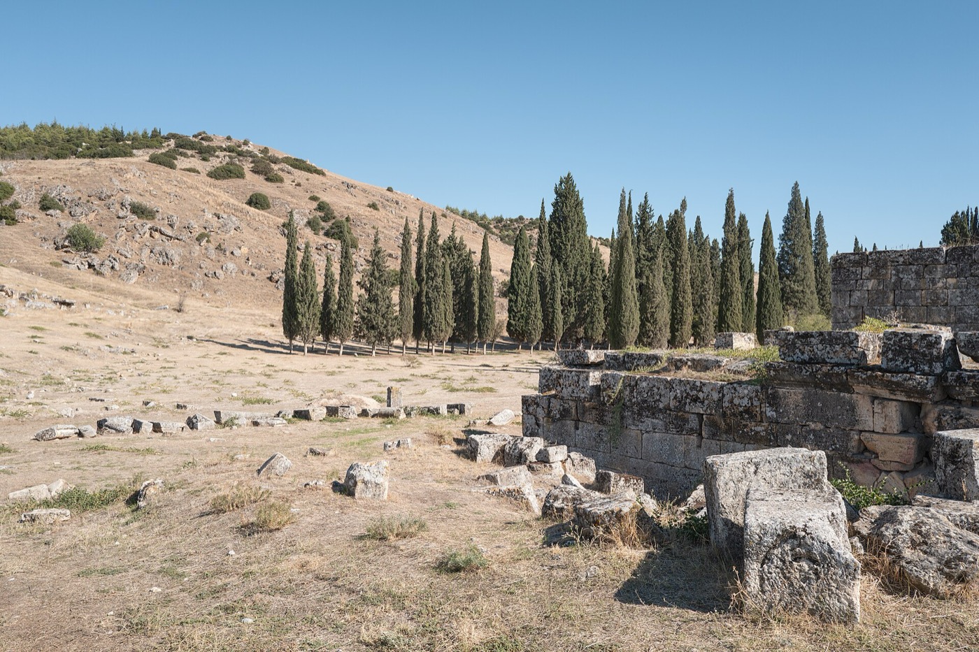 Eine gemauerte Wand der antiken Stadt Hierapolis, dahinter eine Reihe Zypressen am Hang