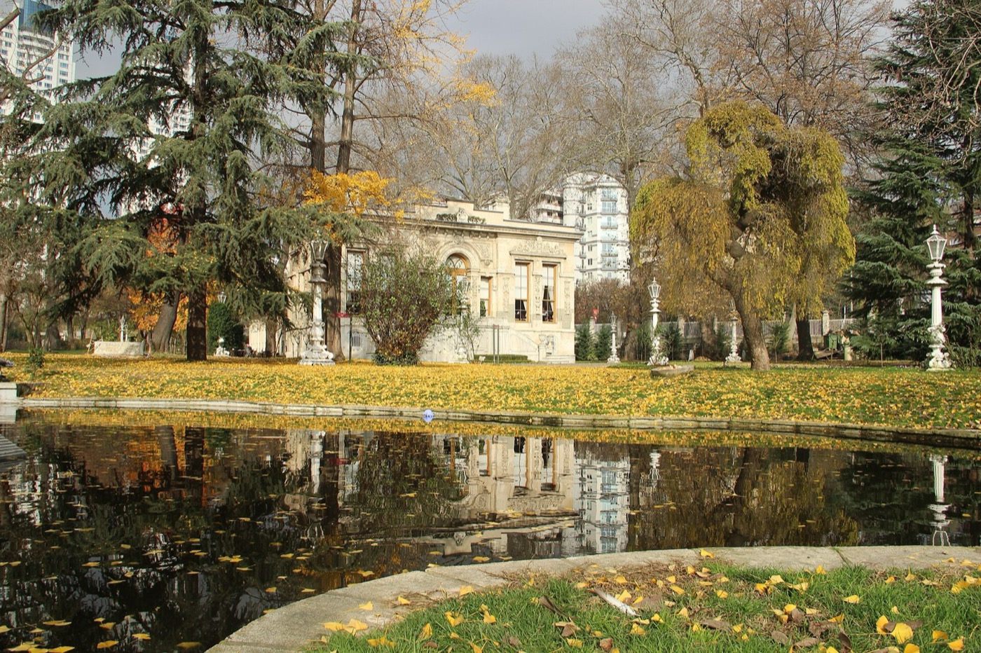 The garden pool at Ihlamur in autumn, with one of the pavilions reflected in the water behind cedars and lamp posts, and yellow leaves on the lawn