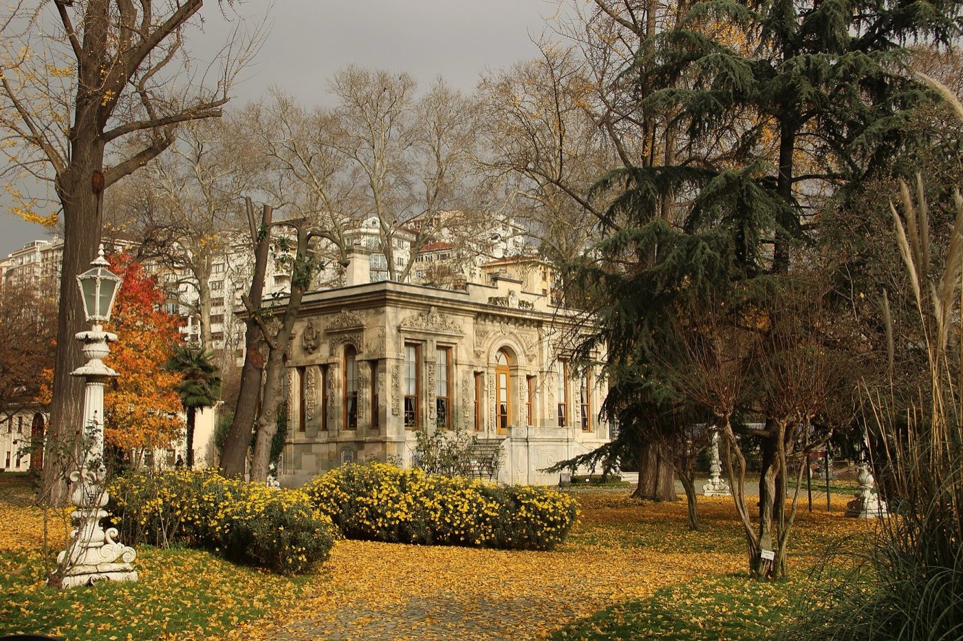 One of the Ihlamur pavilions seen across the garden in autumn, with fallen yellow leaves, a lamp post and shrubs