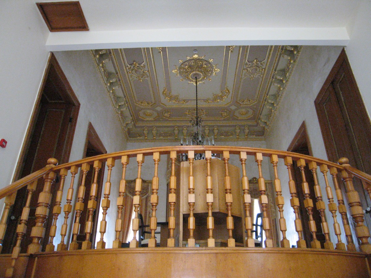Inside the Retinue Pavilion at Ihlamur: a curved wooden balustrade around the upper landing, under a white ceiling with gilded mouldings and a small chandelier