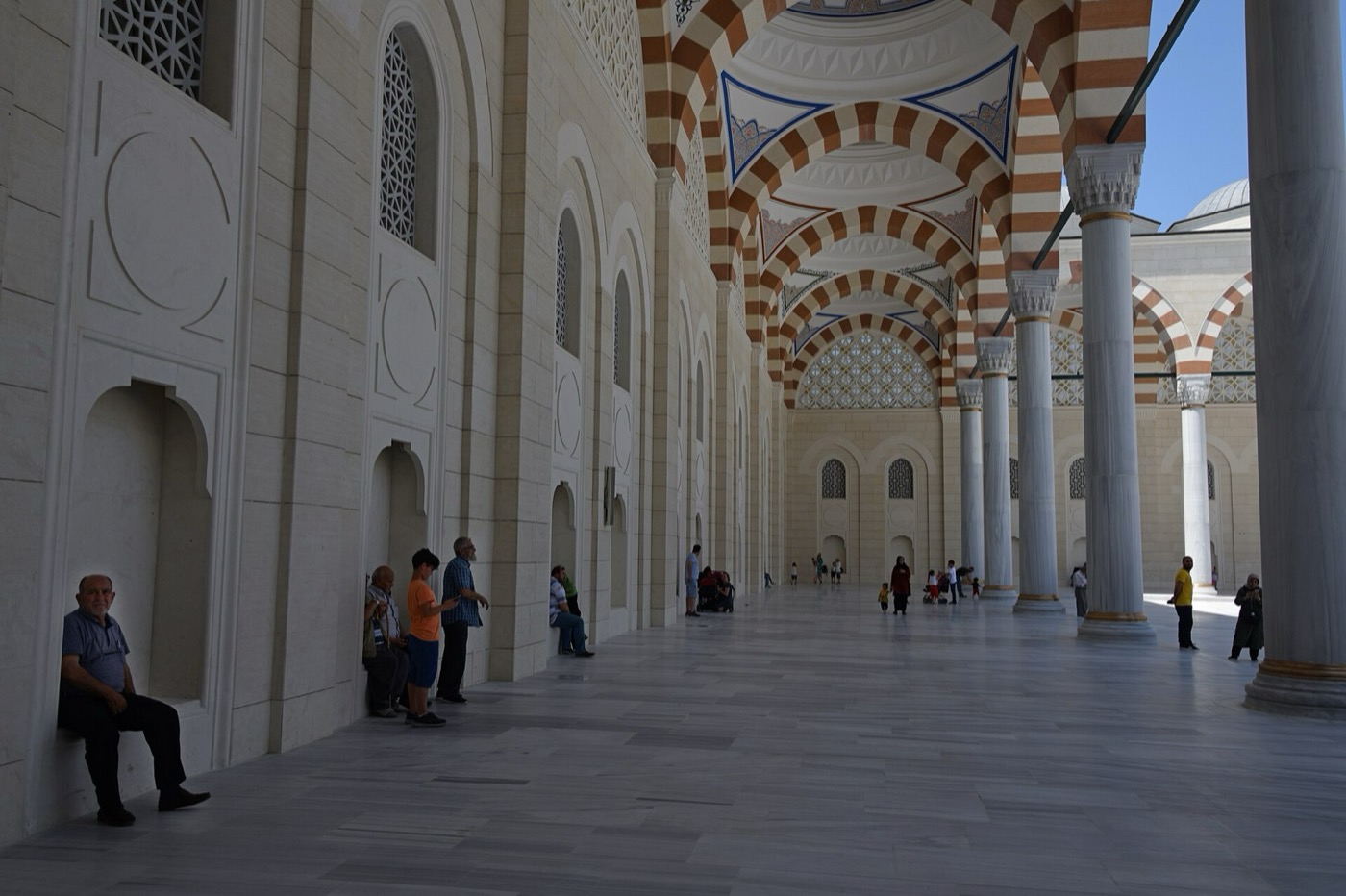 Visitors resting along the shaded arcade on the south-west side of the Büyük Çamlıca courtyard, under red-and-white striped arches