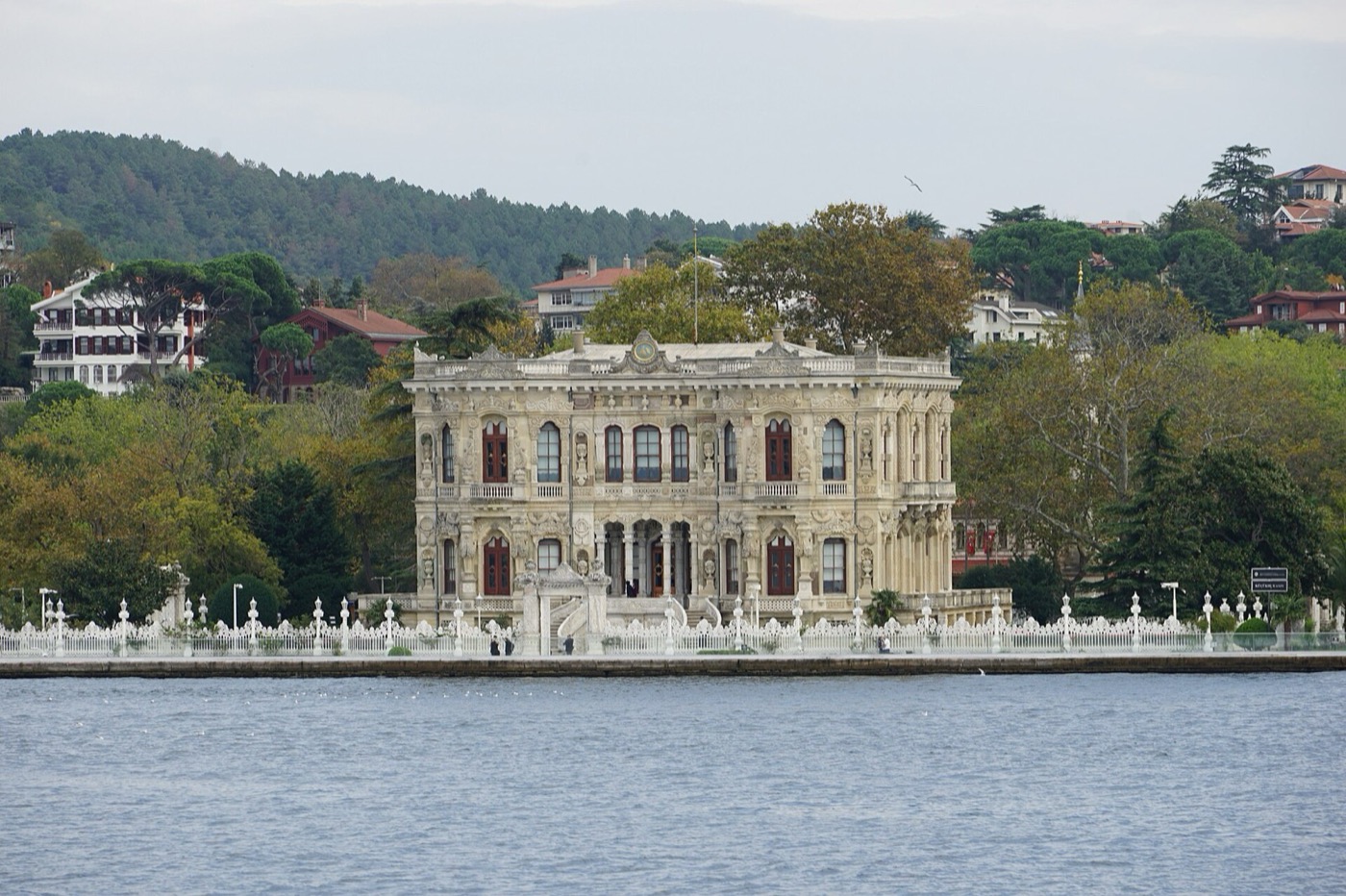Küçüksu Pavilion seen from the Bosphorus: the carved stone facade behind a white openwork railing along the water, with wooded hills and houses behind it