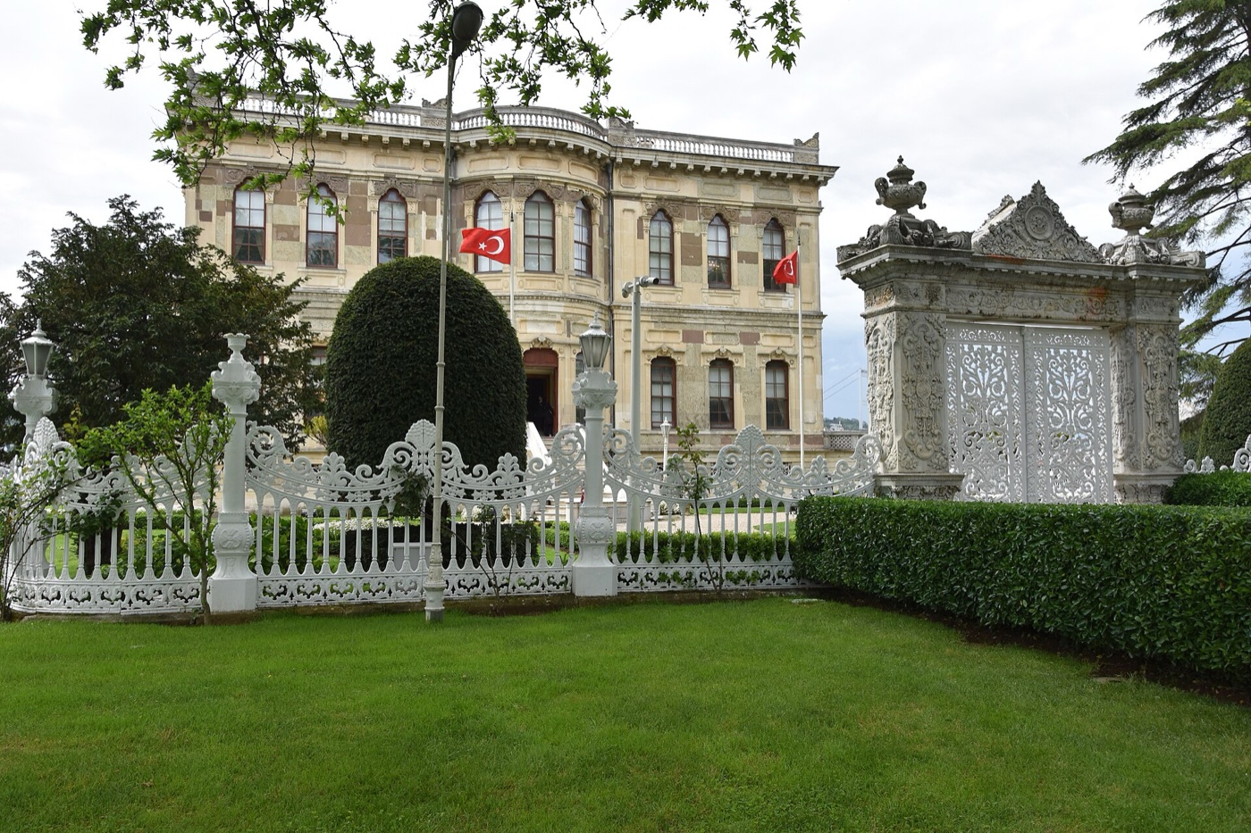 Küçüksu Pavilion behind a white cast-iron railing, with a carved marble gate with latticed doors on the right and a lawn in front