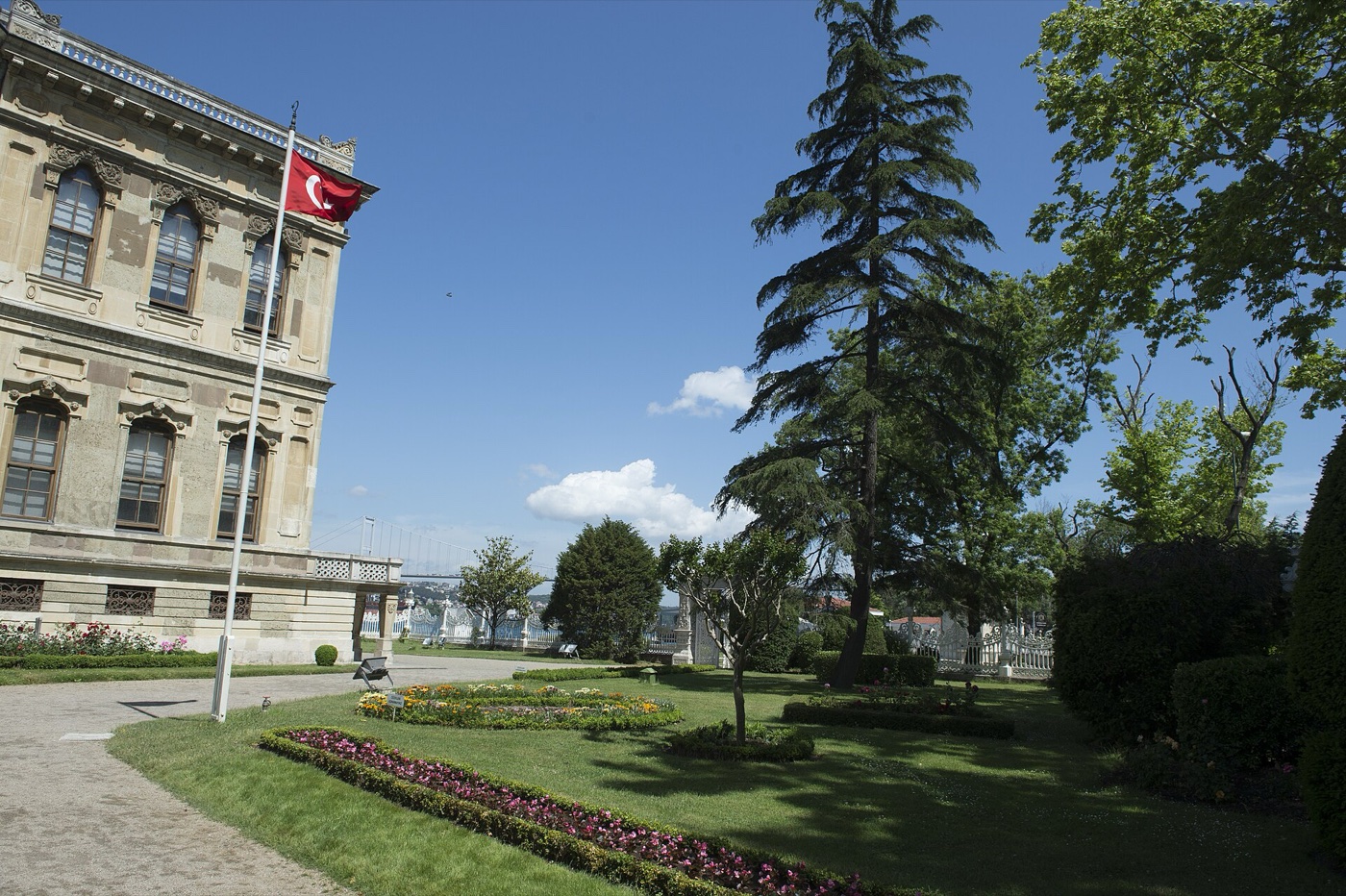 The garden beside Küçüksu Pavilion, with flower beds, tall conifers and a Bosphorus bridge in the distance beyond the waterfront railing