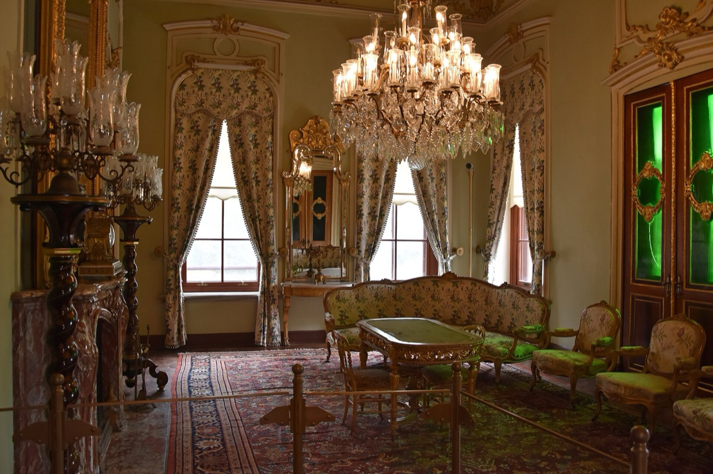 A salon inside Küçüksu Pavilion: a crystal chandelier, floral-upholstered sofa and armchairs around a table, tall curtained windows and a door with green glass panels