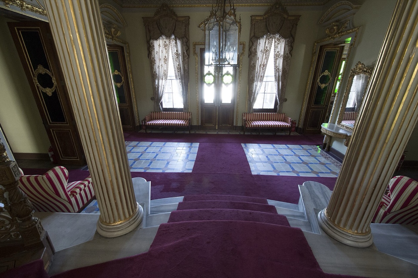 The central hall of Küçüksu Pavilion seen from the top of a carpeted staircase between two gilded columns, with striped benches under the windows