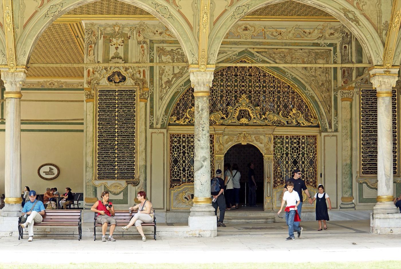 The portico of the Imperial Council building at Topkapı Palace, with marble columns, painted arches and a gilded grille over the entrance, and visitors sitting on benches