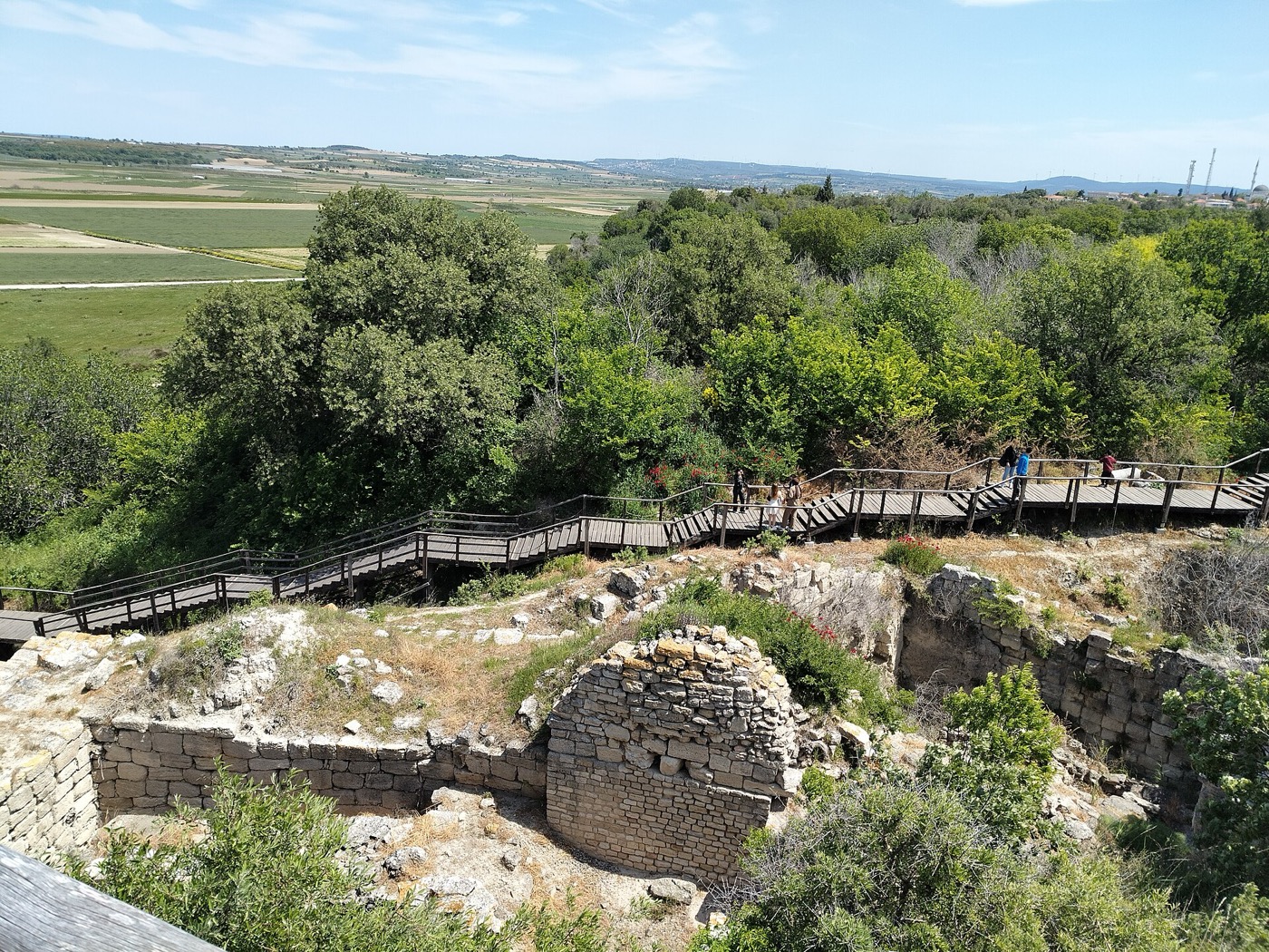 A wooden visitor walkway crossing the excavated ruins of Troy, with the Troad plain beyond