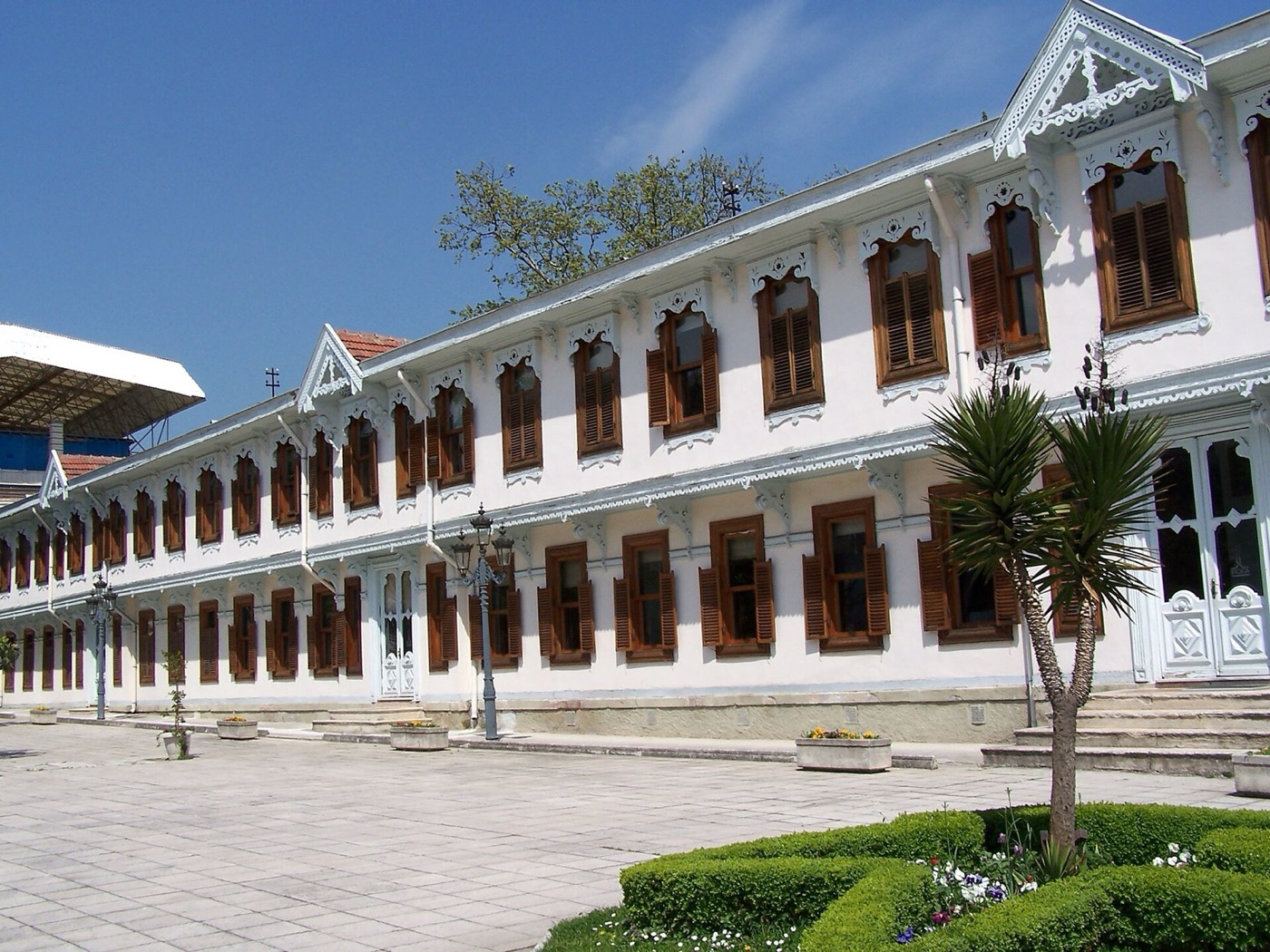 A long two-storey building at Yıldız Palace with white walls, brown shutters and carved wooden trim, facing a paved courtyard with a palm and clipped hedges