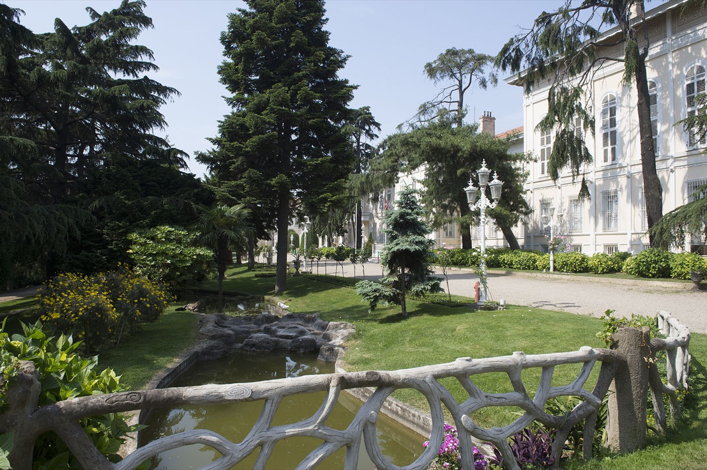 A garden in the Yıldız Palace grounds near the Şale Pavilion: a small pond behind a railing made to look like tree branches, lawns and tall conifers