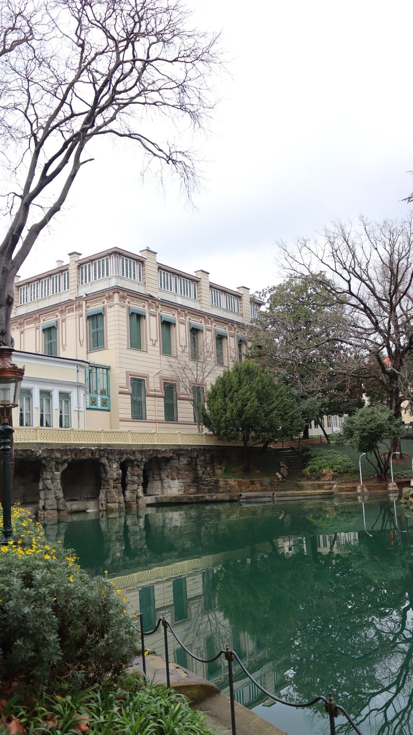 A green pool in the grounds of Yıldız Palace, edged on one side with rock-work grottoes, with a pastel three-storey building and bare winter trees beyond